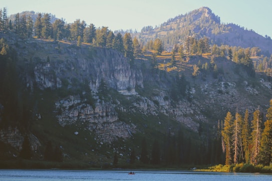 A serene lake bordered by a dense forest of tall evergreen trees, set against the backdrop of rugged, forest-covered mountains. The sunlight gently illuminates the treetops, creating an interplay of light and shadow across the landscape. A lone kayaker paddles quietly across the water, adding a sense of adventure and solitude.