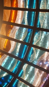 Curved panels of stained glass in various hues, including blue, orange, and clear, create a wavy pattern. The glass appears textured, with some panels having a bubbled or cracked appearance. Strong lighting enhances the vibrant colors and highlights the intricate details of the design.