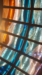 Curved panels of stained glass in various hues, including blue, orange, and clear, create a wavy pattern. The glass appears textured, with some panels having a bubbled or cracked appearance. Strong lighting enhances the vibrant colors and highlights the intricate details of the design.