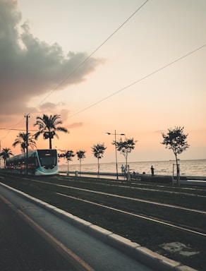 A tram travels along a coastal road lined with palm trees on a track beside the sea. The sky is painted with hues of orange and pink, indicating a sunset. A few silhouetted figures are visible near the waterfront.