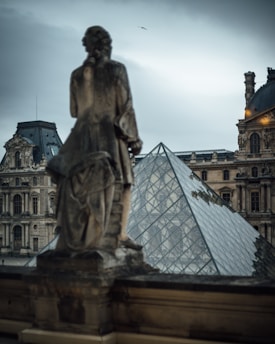 A stone statue stands in the foreground, overlooking the iconic glass pyramid structure of the Louvre Museum with ornate, historic buildings in the background. The sky is overcast, and a bird flies overhead, adding to the atmospheric scene.