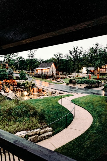 A serene outdoor scene featuring a gently curving paved pathway that leads through well-manicured green lawns. There is a rustic wooden barn illuminated with warm lights, adding a cozy touch. A small waterfall cascades into a pond surrounded by rocks and vibrant flowers, contributing to a tranquil atmosphere. Tall trees frame the background, and there is a soft glow from nearby lights.
