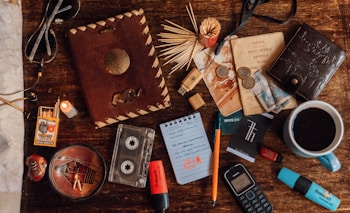 A collection of vintage and personal items laid out on a wooden surface, including a leather-bound notebook with an elephant design, a small notebook with a pen, a pair of sunglasses, earphones, matches, a candle, cassette tapes, foreign currency, an old mobile phone, a USB drive, and a cup of black coffee.
