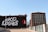 A mural on the side of a building displays the words '#KC Loves' in bold white letters against a black background. Next to the text, there is a circular red emblem with a white rocket illustration. The mural is situated in an urban environment with a clear blue sky above. Adjacent to this building, there is a large red-brick structure with numerous windows.