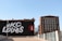 A mural on the side of a building displays the words '#KC Loves' in bold white letters against a black background. Next to the text, there is a circular red emblem with a white rocket illustration. The mural is situated in an urban environment with a clear blue sky above. Adjacent to this building, there is a large red-brick structure with numerous windows.