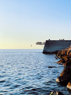 A person is captured mid-air diving from a high platform into the sea. The platform is crowded with spectators watching the dive. The ocean stretches out beneath a clear sky, and a rocky shoreline is visible on the right.