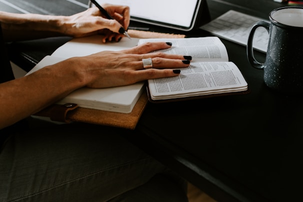 A person writes in a notebook, holding a pen with black painted nails. An open book is nearby, and a mug sits on the dark surface. The setting suggests a workspace or study environment.