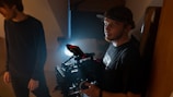 A person holding a professional camera rig, illuminated by a bright light behind them, stands in a dimly lit space. Another person is partially visible to the left, turned slightly away from the camera.