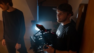 A person holding a professional camera rig, illuminated by a bright light behind them, stands in a dimly lit space. Another person is partially visible to the left, turned slightly away from the camera.