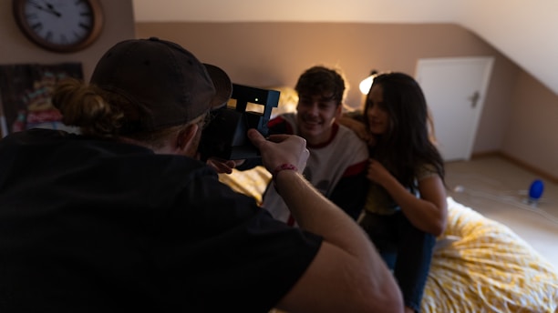 A person in a cap is holding a camera, taking a picture of a couple sitting together on a bed in a cozy room. The room has warm lighting with a clock on the wall and a closed door in the background. The atmosphere appears relaxed and casual.