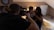 A person in a cap is holding a camera, taking a picture of a couple sitting together on a bed in a cozy room. The room has warm lighting with a clock on the wall and a closed door in the background. The atmosphere appears relaxed and casual.
