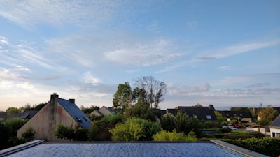 A peaceful residential neighborhood featuring a few houses with sloped roofs and surrounding greenery, set against a backdrop of a bright blue sky with scattered white clouds. The area is lush with various trees and bushes.