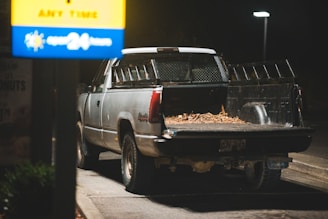 A pickup truck parked at night under street lighting, loaded with leaves or debris in its open cargo area. A partially visible sign with a yellow background is on the left.