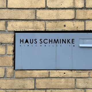 A gray metal sign with cut-out black letters displays the words 'Haus Schminke' and 'Kirschallee 1b.' It's mounted on a background of tan brick wall, providing a stark contrast between the modern sign and the traditional brick texture.