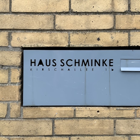 A gray metal sign with cut-out black letters displays the words 'Haus Schminke' and 'Kirschallee 1b.' It's mounted on a background of tan brick wall, providing a stark contrast between the modern sign and the traditional brick texture.