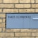A gray metal sign with cut-out black letters displays the words 'Haus Schminke' and 'Kirschallee 1b.' It's mounted on a background of tan brick wall, providing a stark contrast between the modern sign and the traditional brick texture.