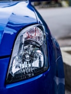 A close-up view of the headlight of a blue car. The headlight has a clear lens and intricate details of the reflector and bulb inside. The car's body paint is glossy and reflects the surrounding environment.