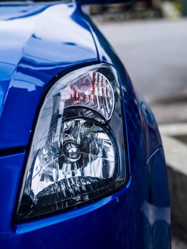 A close-up view of the headlight of a blue car. The headlight has a clear lens and intricate details of the reflector and bulb inside. The car's body paint is glossy and reflects the surrounding environment.