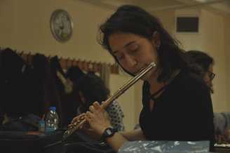 A woman is playing a flute while seated indoors, with closed eyes and an expression of concentration. Behind her, other people are present, and coats hang from hooks along the wall. A clock is visible on the wall, indicating it is a room designed for group activities or study.