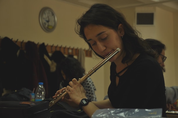 A woman is playing a flute while seated indoors, with closed eyes and an expression of concentration. Behind her, other people are present, and coats hang from hooks along the wall. A clock is visible on the wall, indicating it is a room designed for group activities or study.