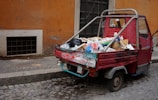A small, red three-wheeled vehicle loaded with various types of garbage, including cardboard boxes, plastic bags, and miscellaneous waste, is parked on a cobblestone street. The vehicle is adjacent to a weathered, orange-toned wall and a black door. The scene suggests an urban environment, possibly in need of cleaning or waste management.