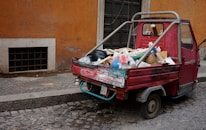 A small, red three-wheeled vehicle loaded with various types of garbage, including cardboard boxes, plastic bags, and miscellaneous waste, is parked on a cobblestone street. The vehicle is adjacent to a weathered, orange-toned wall and a black door. The scene suggests an urban environment, possibly in need of cleaning or waste management.
