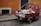 A small, red three-wheeled vehicle loaded with various types of garbage, including cardboard boxes, plastic bags, and miscellaneous waste, is parked on a cobblestone street. The vehicle is adjacent to a weathered, orange-toned wall and a black door. The scene suggests an urban environment, possibly in need of cleaning or waste management.