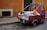 A small, red three-wheeled vehicle loaded with various types of garbage, including cardboard boxes, plastic bags, and miscellaneous waste, is parked on a cobblestone street. The vehicle is adjacent to a weathered, orange-toned wall and a black door. The scene suggests an urban environment, possibly in need of cleaning or waste management.