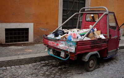 A small, red three-wheeled vehicle loaded with various types of garbage, including cardboard boxes, plastic bags, and miscellaneous waste, is parked on a cobblestone street. The vehicle is adjacent to a weathered, orange-toned wall and a black door. The scene suggests an urban environment, possibly in need of cleaning or waste management.