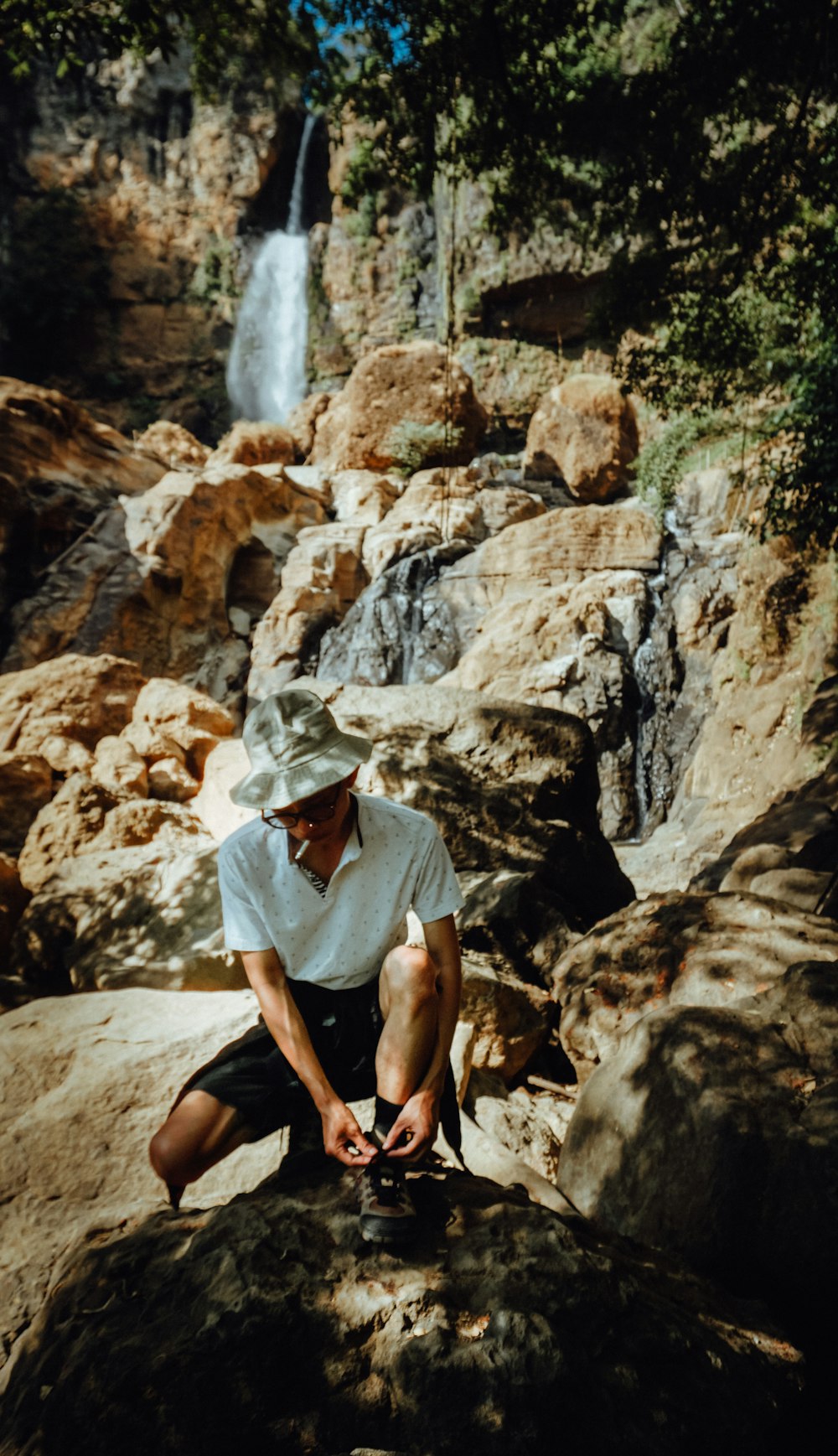 Frau im weißen Hijab sitzt tagsüber auf braunem Felsen in der Nähe von Wasserfällen