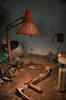A dimly lit workshop features various tools scattered across a well-worn table. A large, rust-colored metal lamp hangs overhead, casting a warm glow. On the table, there are items including a red-handled multitool, a rusty metal bracket, a ratchet wrench, and a spray can.