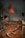 A dimly lit workshop features various tools scattered across a well-worn table. A large, rust-colored metal lamp hangs overhead, casting a warm glow. On the table, there are items including a red-handled multitool, a rusty metal bracket, a ratchet wrench, and a spray can.