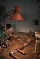 A dimly lit workshop features various tools scattered across a well-worn table. A large, rust-colored metal lamp hangs overhead, casting a warm glow. On the table, there are items including a red-handled multitool, a rusty metal bracket, a ratchet wrench, and a spray can.