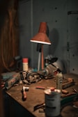 A dimly lit workshop table scattered with various tools and equipment, including a drill, a hammer, aerosol cans, a utility knife, and a red lamp casting a warm glow on the surface. The setting appears disorganized, with dust and clutter indicative of a working environment.