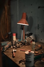 A dimly lit workshop table scattered with various tools and equipment, including a drill, a hammer, aerosol cans, a utility knife, and a red lamp casting a warm glow on the surface. The setting appears disorganized, with dust and clutter indicative of a working environment.