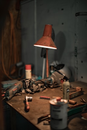 A dimly lit workshop table scattered with various tools and equipment, including a drill, a hammer, aerosol cans, a utility knife, and a red lamp casting a warm glow on the surface. The setting appears disorganized, with dust and clutter indicative of a working environment.