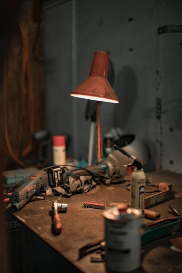 A dimly lit workshop table scattered with various tools and equipment, including a drill, a hammer, aerosol cans, a utility knife, and a red lamp casting a warm glow on the surface. The setting appears disorganized, with dust and clutter indicative of a working environment.