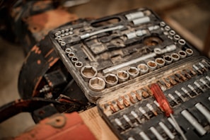 A well-used toolset with various socket wrenches and sockets organized within a rectangular black plastic case. A red-handled screwdriver is placed among the metal tools, which show signs of wear and rusting. The surface underneath is blurred and appears to be wooden, with parts of a dark metallic object in the background.