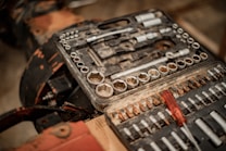 A well-used toolset with various socket wrenches and sockets organized within a rectangular black plastic case. A red-handled screwdriver is placed among the metal tools, which show signs of wear and rusting. The surface underneath is blurred and appears to be wooden, with parts of a dark metallic object in the background.