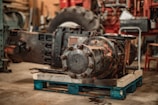A large industrial machine component is placed on a wooden pallet in a workshop setting. The component is made of metal with visible rust spots, and bolts are holding its circular sections together. The surrounding environment includes a large tire, suggesting a heavy-duty machinery context, tools, and equipment in the background, with tones of brown and red dominating the scene.