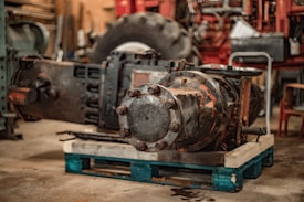 A large industrial machine component is placed on a wooden pallet in a workshop setting. The component is made of metal with visible rust spots, and bolts are holding its circular sections together. The surrounding environment includes a large tire, suggesting a heavy-duty machinery context, tools, and equipment in the background, with tones of brown and red dominating the scene.