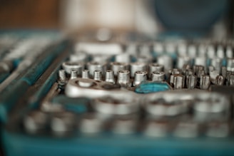 A close-up of a set of metallic socket wrenches and other related tools, arranged in an orderly manner. The tools have a silver finish and are housed in a blue tray or case, partially blurred.