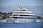 A large, luxurious yacht is anchored on a calm body of water near a coastal area with lush greenery and distant mountains. The yacht has multiple decks and modern design features, with people seen relaxing on the lower deck. The background showcases a serene natural landscape with a clear blue sky.