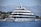 A large, luxurious yacht is anchored on a calm body of water near a coastal area with lush greenery and distant mountains. The yacht has multiple decks and modern design features, with people seen relaxing on the lower deck. The background showcases a serene natural landscape with a clear blue sky.