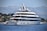 A large, luxurious yacht is anchored on a calm body of water near a coastal area with lush greenery and distant mountains. The yacht has multiple decks and modern design features, with people seen relaxing on the lower deck. The background showcases a serene natural landscape with a clear blue sky.