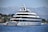 A large, luxurious yacht is anchored on a calm body of water near a coastal area with lush greenery and distant mountains. The yacht has multiple decks and modern design features, with people seen relaxing on the lower deck. The background showcases a serene natural landscape with a clear blue sky.