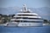 A large, luxurious yacht is anchored on a calm body of water near a coastal area with lush greenery and distant mountains. The yacht has multiple decks and modern design features, with people seen relaxing on the lower deck. The background showcases a serene natural landscape with a clear blue sky.