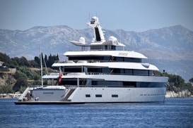 A large, luxurious yacht is anchored on a calm body of water near a coastal area with lush greenery and distant mountains. The yacht has multiple decks and modern design features, with people seen relaxing on the lower deck. The background showcases a serene natural landscape with a clear blue sky.