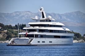 A large, luxurious yacht is anchored on a calm body of water near a coastal area with lush greenery and distant mountains. The yacht has multiple decks and modern design features, with people seen relaxing on the lower deck. The background showcases a serene natural landscape with a clear blue sky.