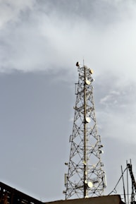 A tall metal communication tower equipped with numerous satellite dishes and antennas stands against a backdrop of a cloudy sky. The structure is intricate, composed of lattice frameworks that provide support and stability.
